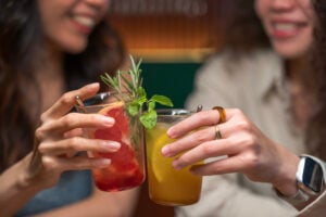 Two young Asian Chinese women, a lesbian couple, toasting colorful cocktails and smiling warmly at each other in a cozy restaurant, celebrating and enjoying the moment, warm lighting and modern interior, Kuala Lumpur, Malaysia
