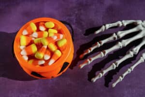 Stock photo showing close-up view of a model, skeletal hand reaching towards a plastic, orange, Halloween trick or treat collection bucket, shaped like a pumpkin Jack O'Lantern, filled with candy corn.